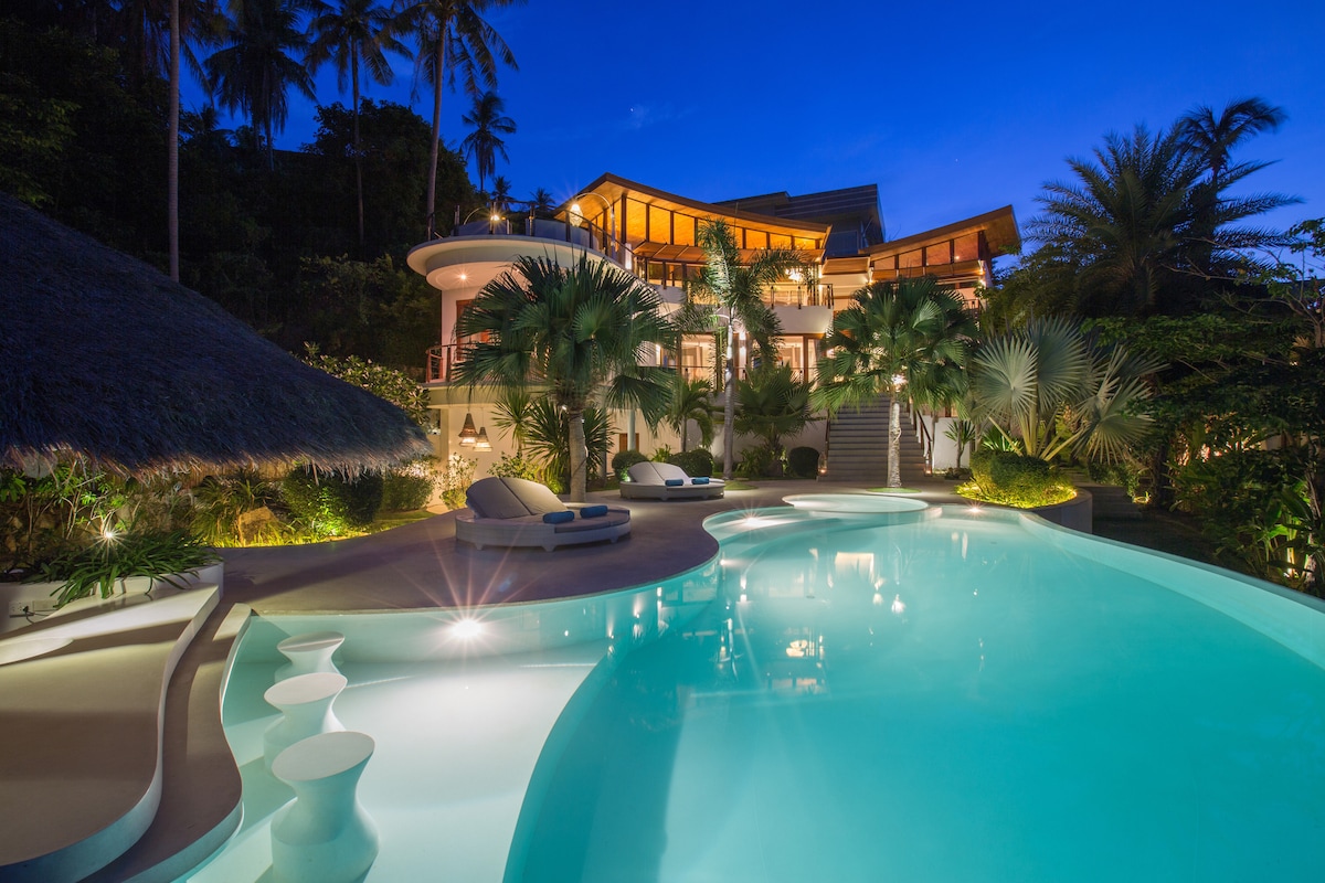 The villa is illuminated at night, showcasing a sparkling swimming pool surrounded by lush tropical landscaping. Modern lounge chairs are positioned near the water's edge, with gentle lighting enhancing the serene atmosphere. The multi-level villa features large windows, providing glimpses of the interior.