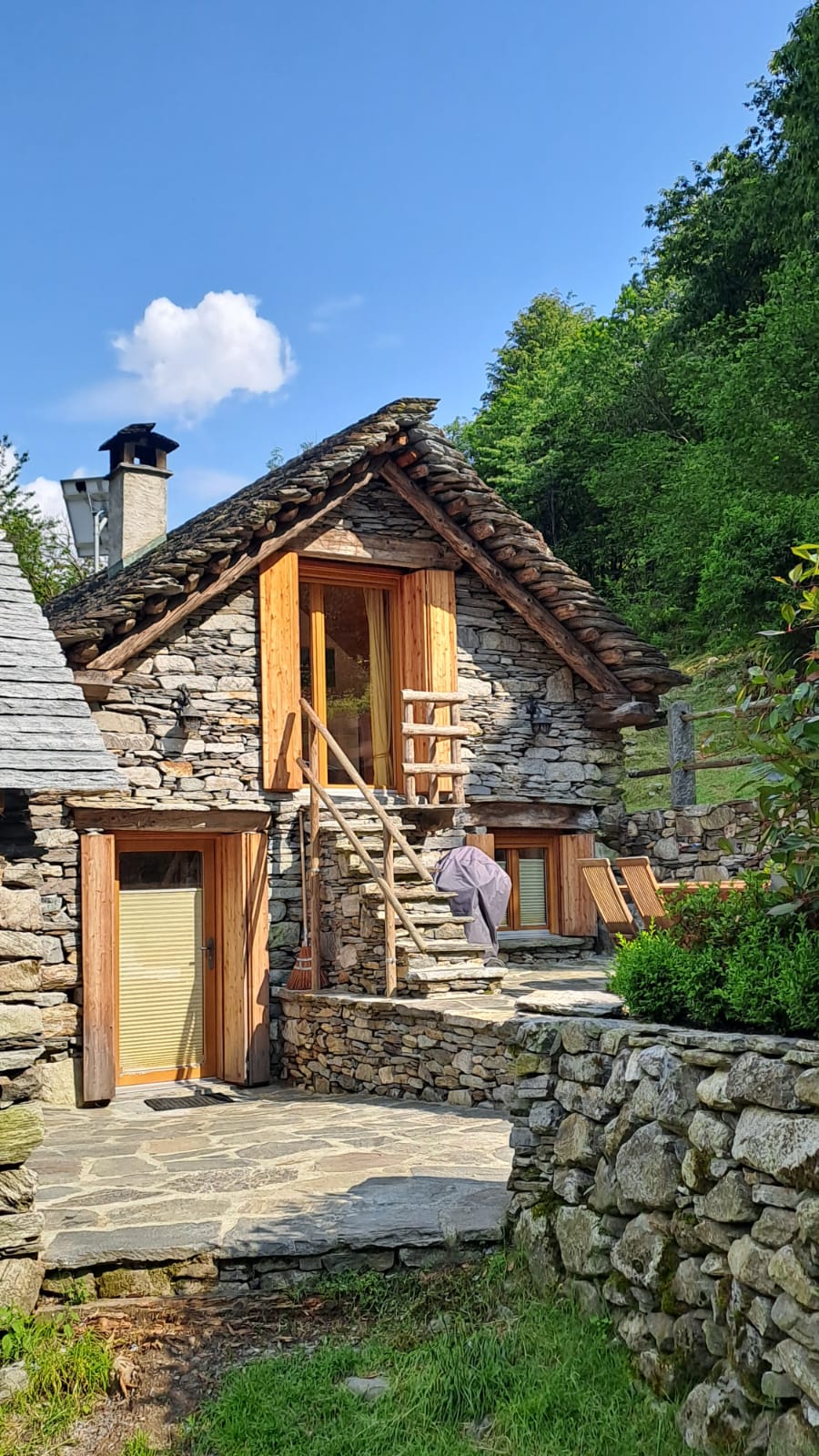 The charming rustic stone house showcases a pitched roof and wooden accents. Natural stone steps lead to the entrance, framed by lush greenery. Sunlight illuminates the façade, highlighting the handcrafted details of the door and windows.