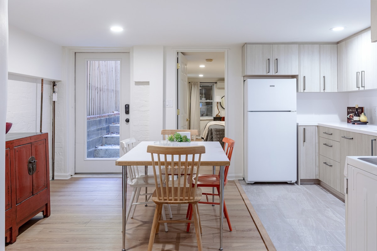 An eat-in kitchen is presented with a wooden dining table surrounded by chairs in yellow and red. A refrigerator and modern cabinetry are visible against a backdrop of light-colored walls. A door leads to the outside, with steps leading up to a patio.