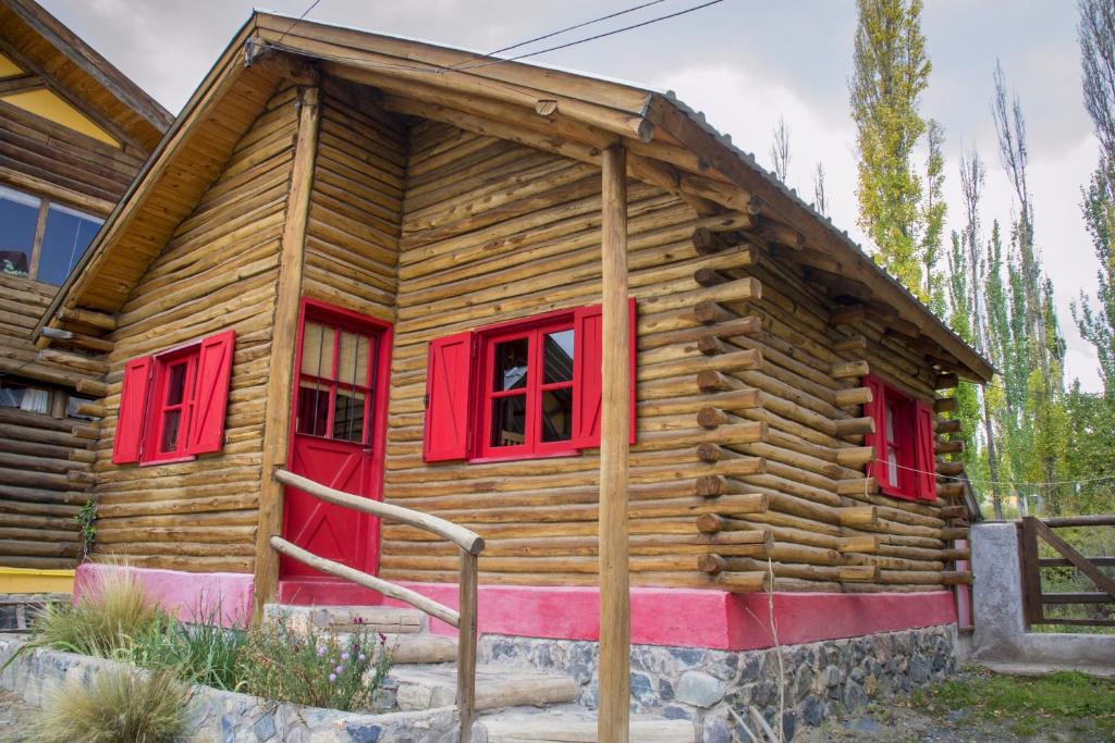 A rustic wooden cabin features distinctive red shutters and a vibrant door. The structure is made of logs, providing a natural appearance. Steps lead up to the entrance, surrounded by low shrubs and stones, emphasizing the cabin's connection to the serene outdoor environment.