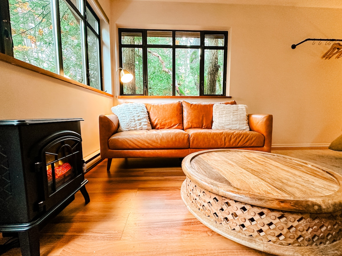 A cozy living area features a brown leather sofa adorned with two soft pillows. A round wooden coffee table sits in front, complementing the warm tones of the space. Natural light streams through large windows, offering views of the surrounding greenery, while an electric fireplace provides warmth.