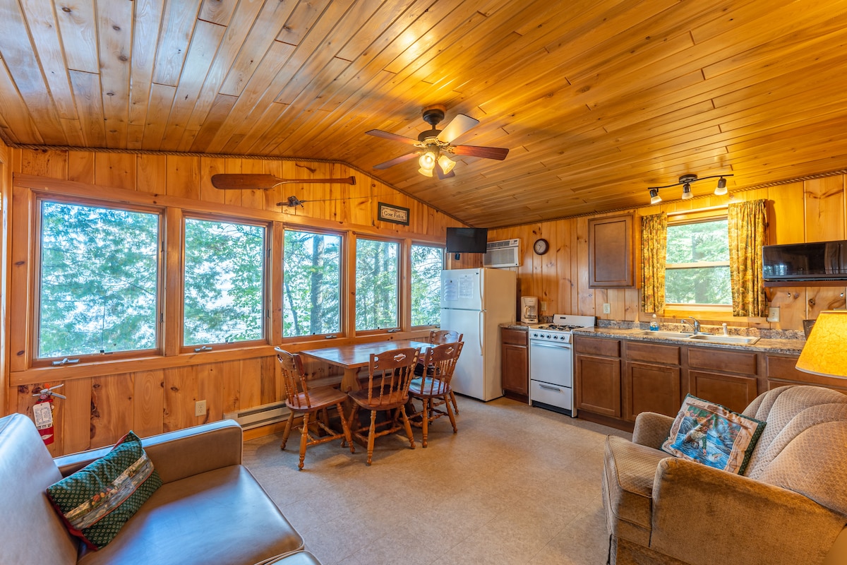 A welcoming cabin interior features wooden walls and a vaulted ceiling. Large windows provide a view of the surrounding trees. A dining table with four chairs is positioned near the kitchen, equipped with modern appliances and warm lighting, while a comfortable seating area includes a sofa.