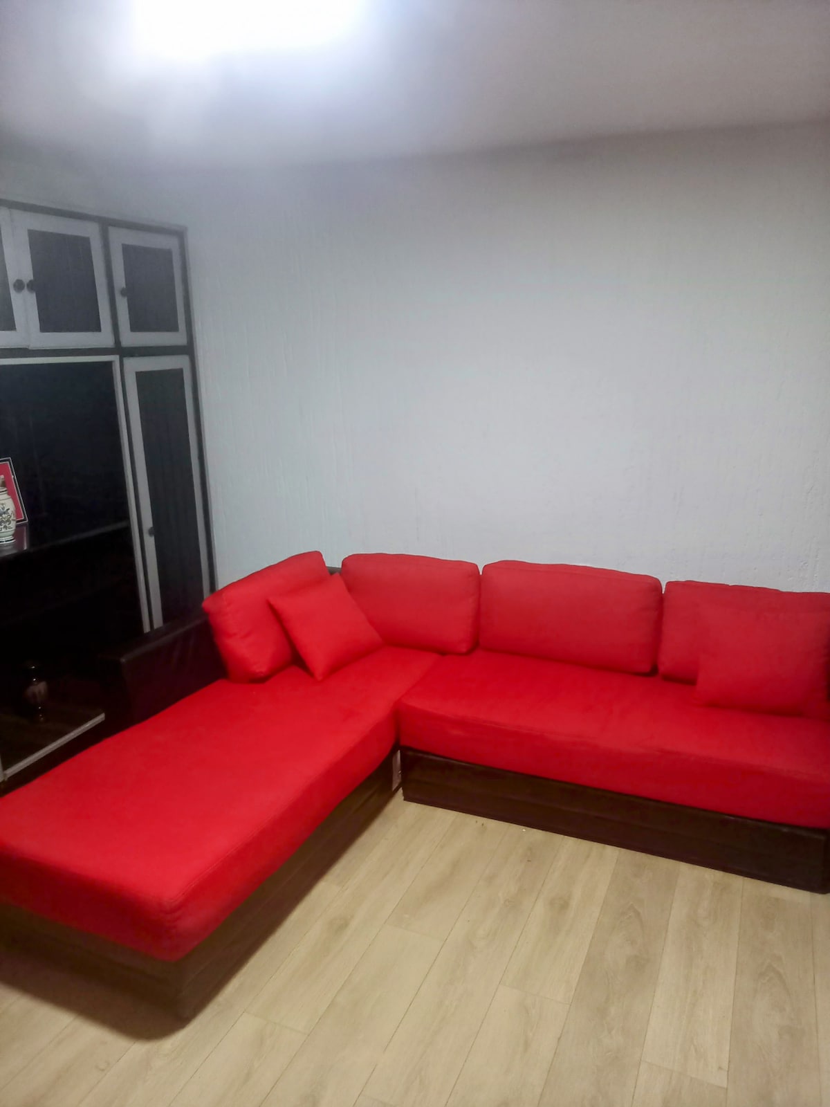 A section of a living area is presented featuring a red sectional sofa that provides ample seating. The sofa is complemented by square cushions. A dark wood shelf is visible in the background, displaying various items, and light-colored flooring enhances the space.
