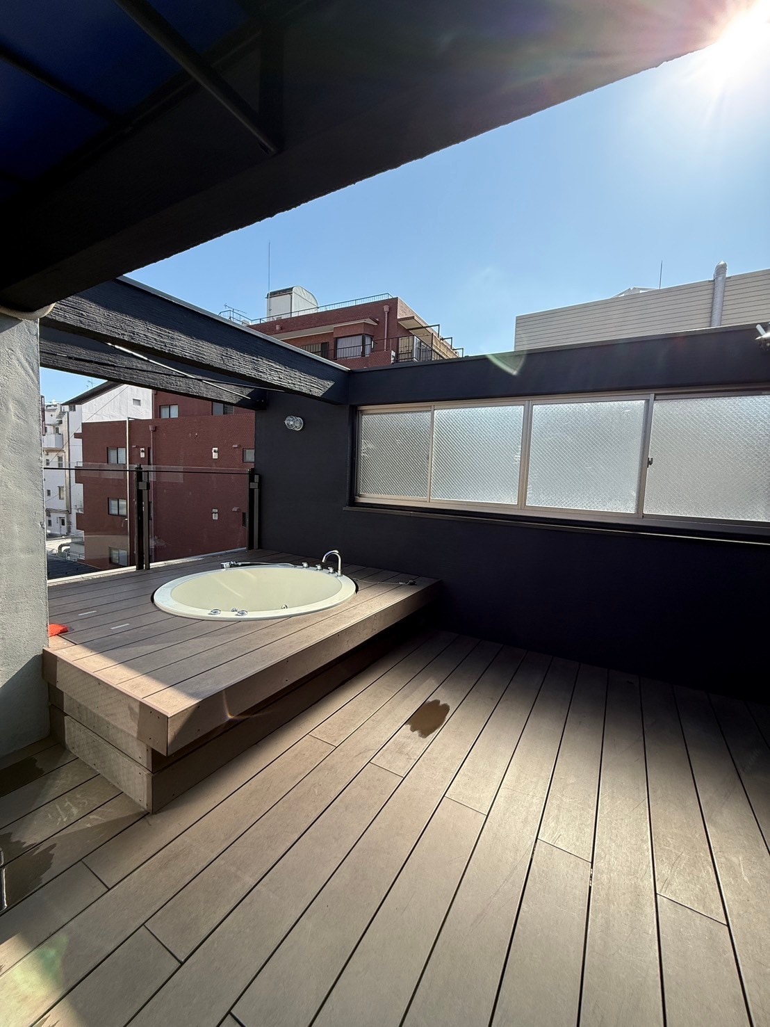 An outdoor space features a circular hot tub set on a wooden platform. Sunlight filters through the glass windows, highlighting the smooth gray walls and warm wooden flooring. The area is surrounded by buildings, offering a private retreat.