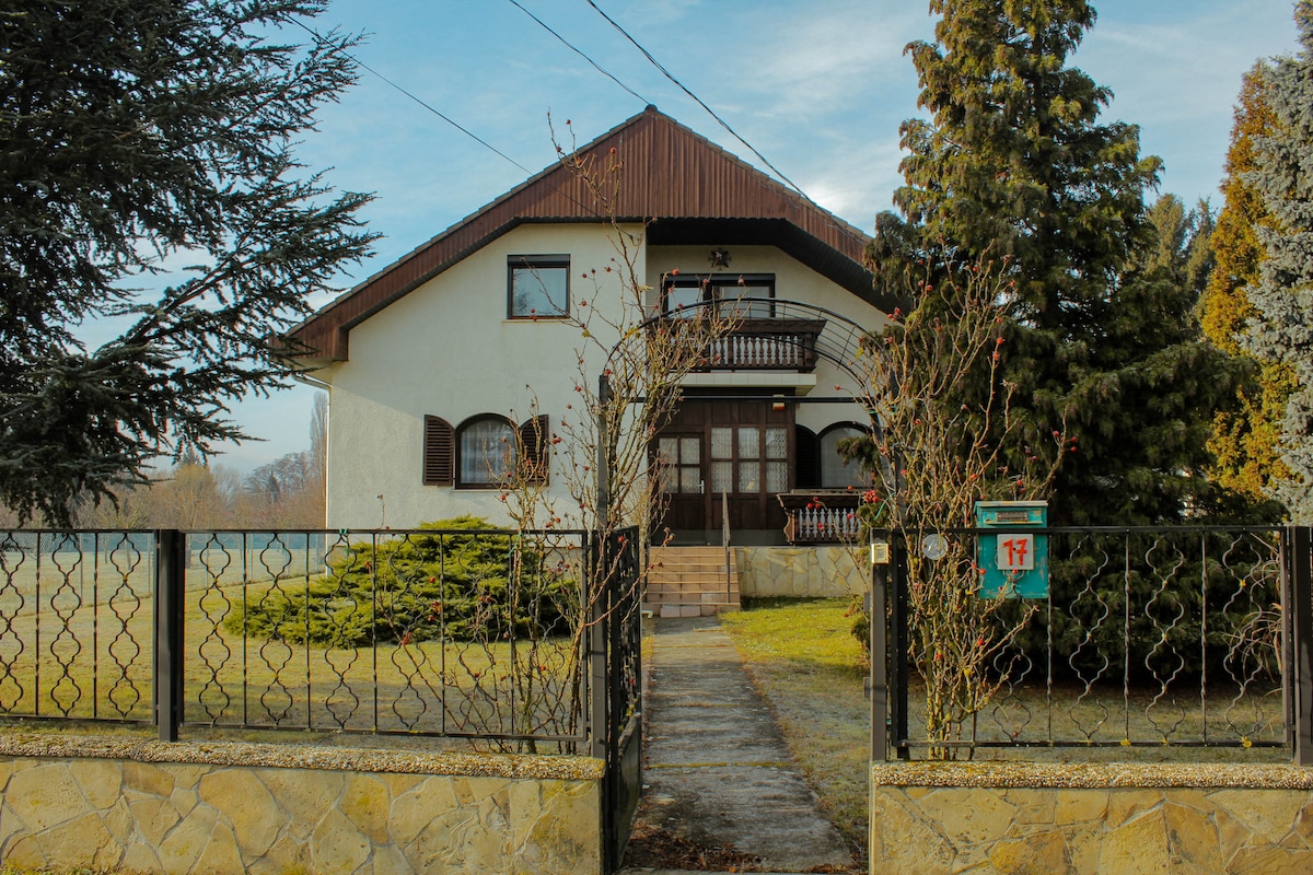 A two-story house with a distinctive design is framed by a well-maintained garden. A paved path leads to the entrance, which is characterized by a covered porch. Tall trees and shrubs surround the property, offering an inviting outdoor setting.