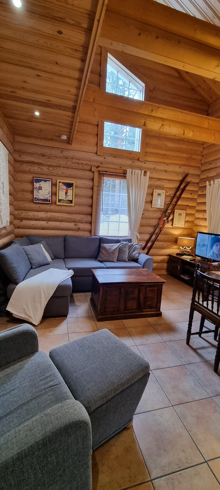 A cozy living area features a large sectional sofa in a neutral tone, complemented by a wooden coffee table. Natural light filters through large windows, illuminating the log walls. A small dining table and entertainment center complete the inviting atmosphere.