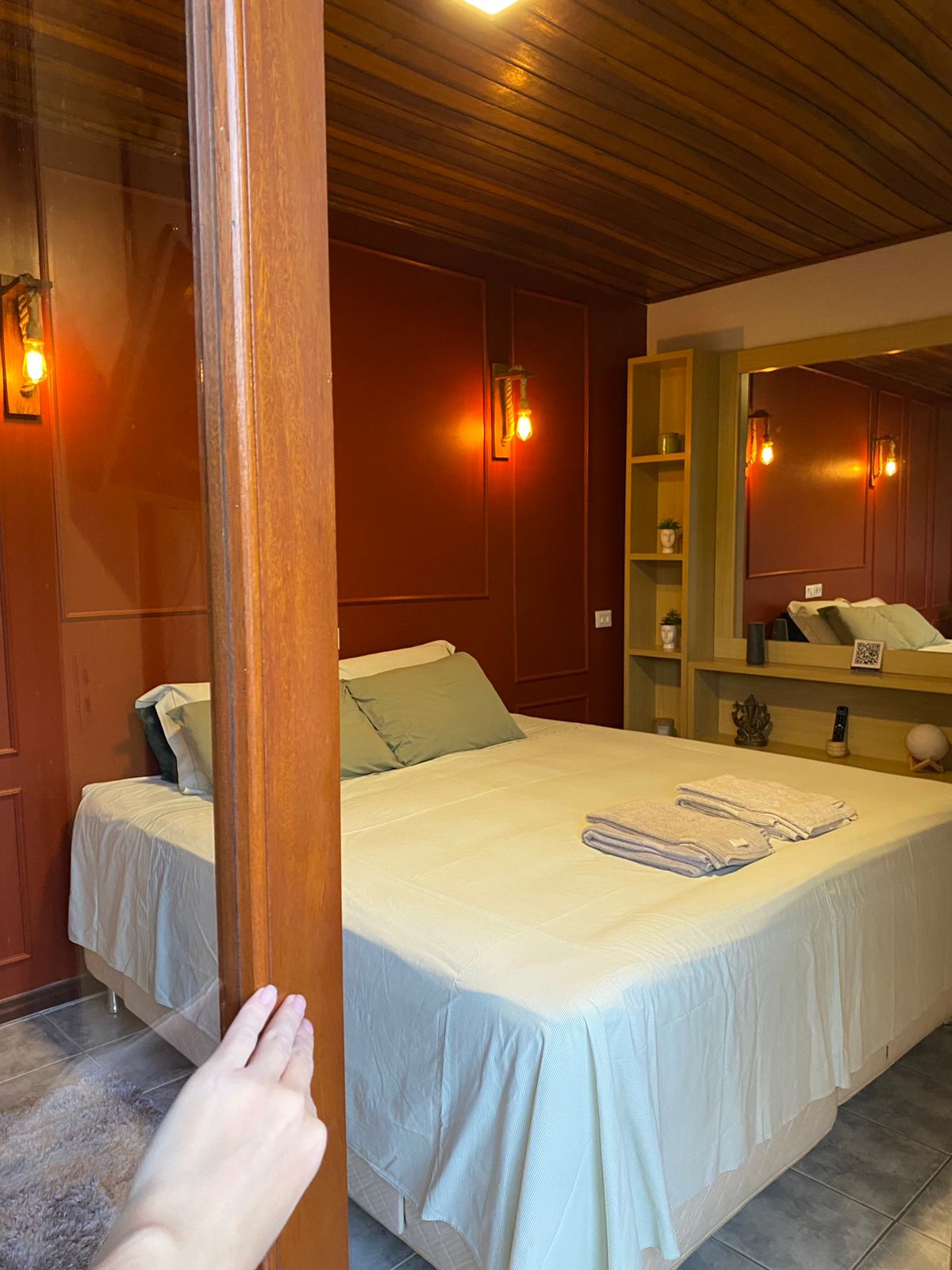 A private room is featured with warm red walls and wooden ceiling. A spacious bed is positioned next to a stylish shelving unit and a mirror. Two light fixtures provide soft illumination, while neatly folded towels are placed on the bed.