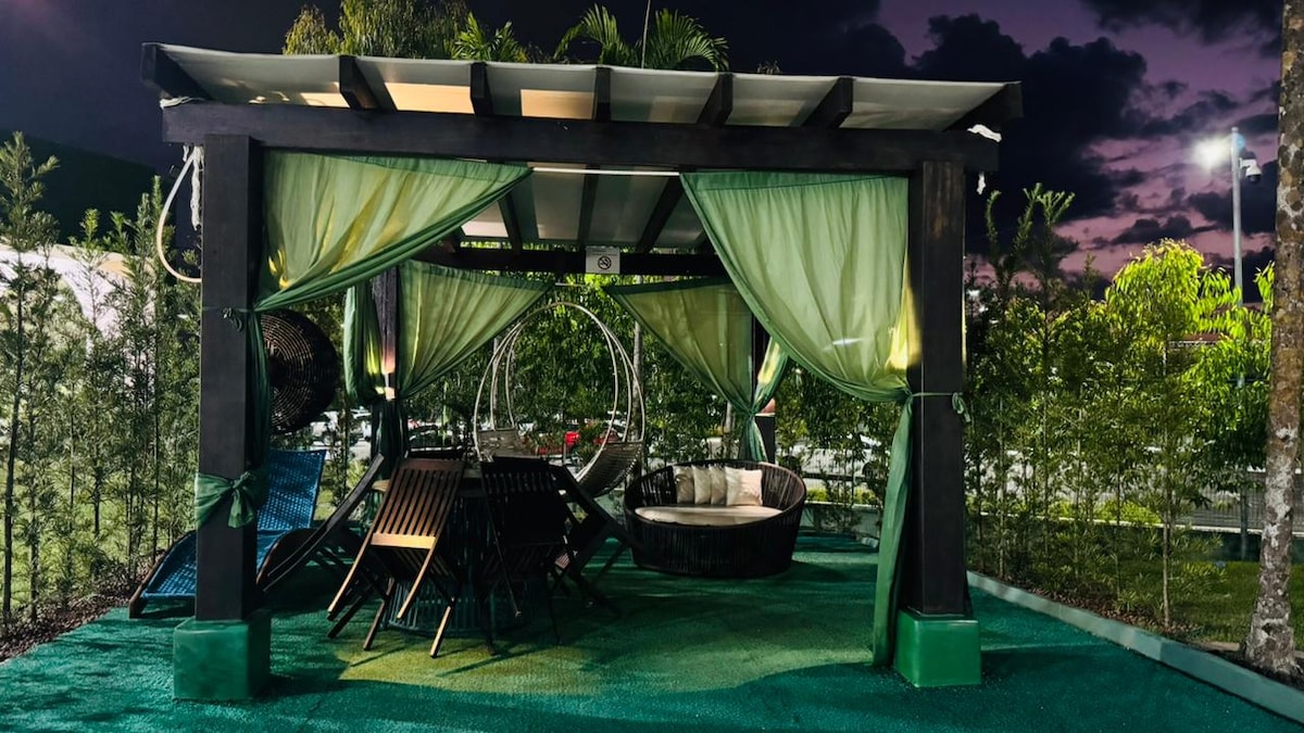 A spacious outdoor seating area is sheltered under a pergola, featuring a circular lounge chair and several stylish loungers. Soft green drapes adorn the structure, creating a relaxing vibe amidst lush greenery. Gentle lighting enhances the evening atmosphere.