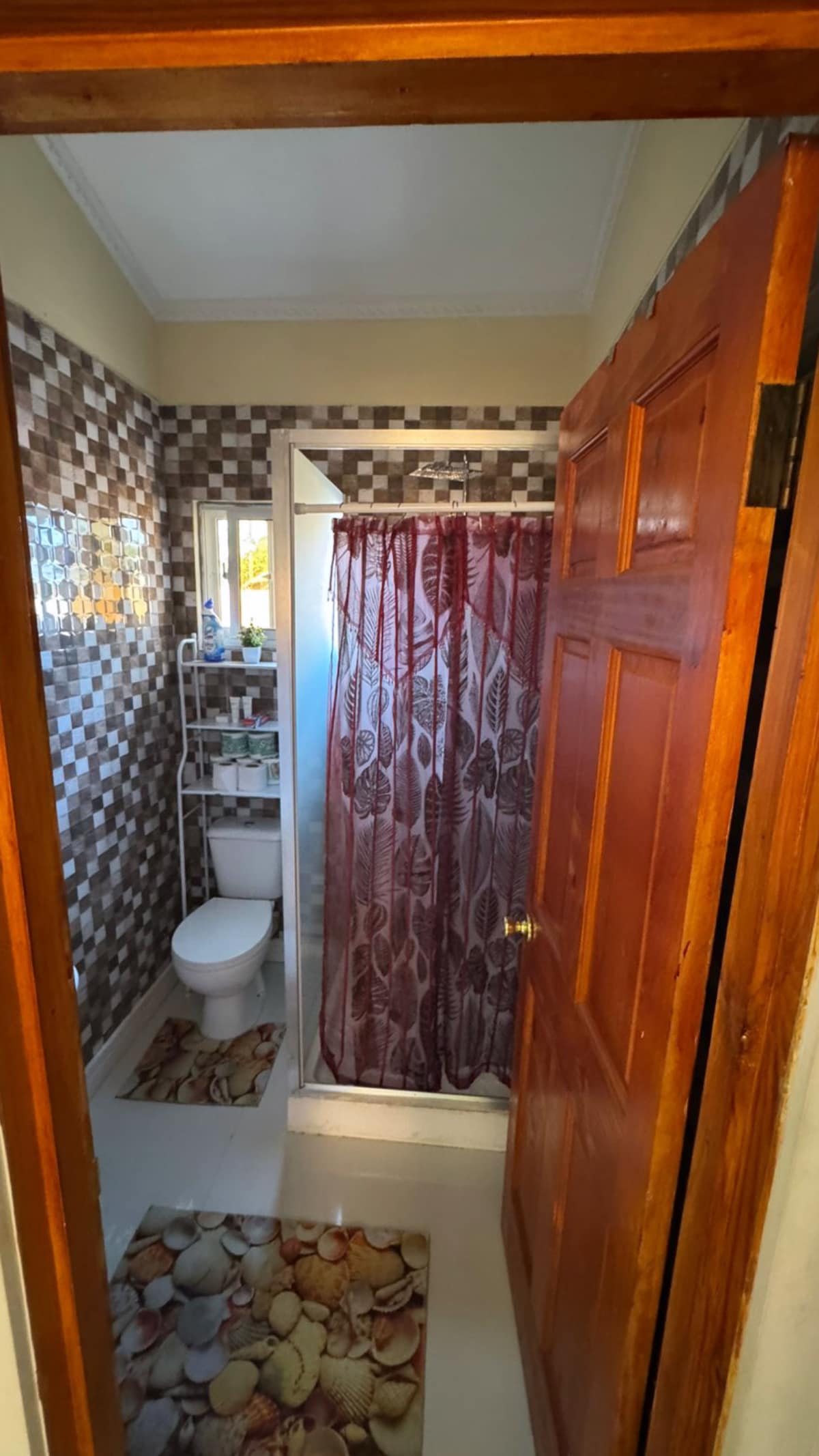 A compact bathroom features a shower enclosed with a patterned curtain, a toilet, and a wall-mounted shelf holding toiletries. The tiled walls are adorned with a mosaic pattern, while a decorative mat with a pebble design lays on the floor, providing a soothing touch to the space.