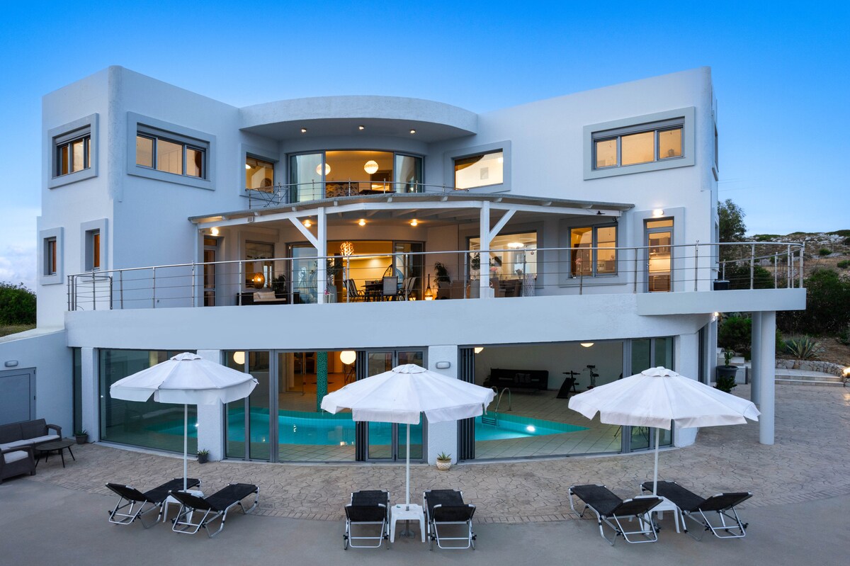 The villa's modern exterior is showcased with large windows and multiple terraces. A spacious patio area features lounge chairs and umbrellas, while the heated indoor pool is visible through the glass doors, creating a seamless connection between indoor and outdoor spaces.
