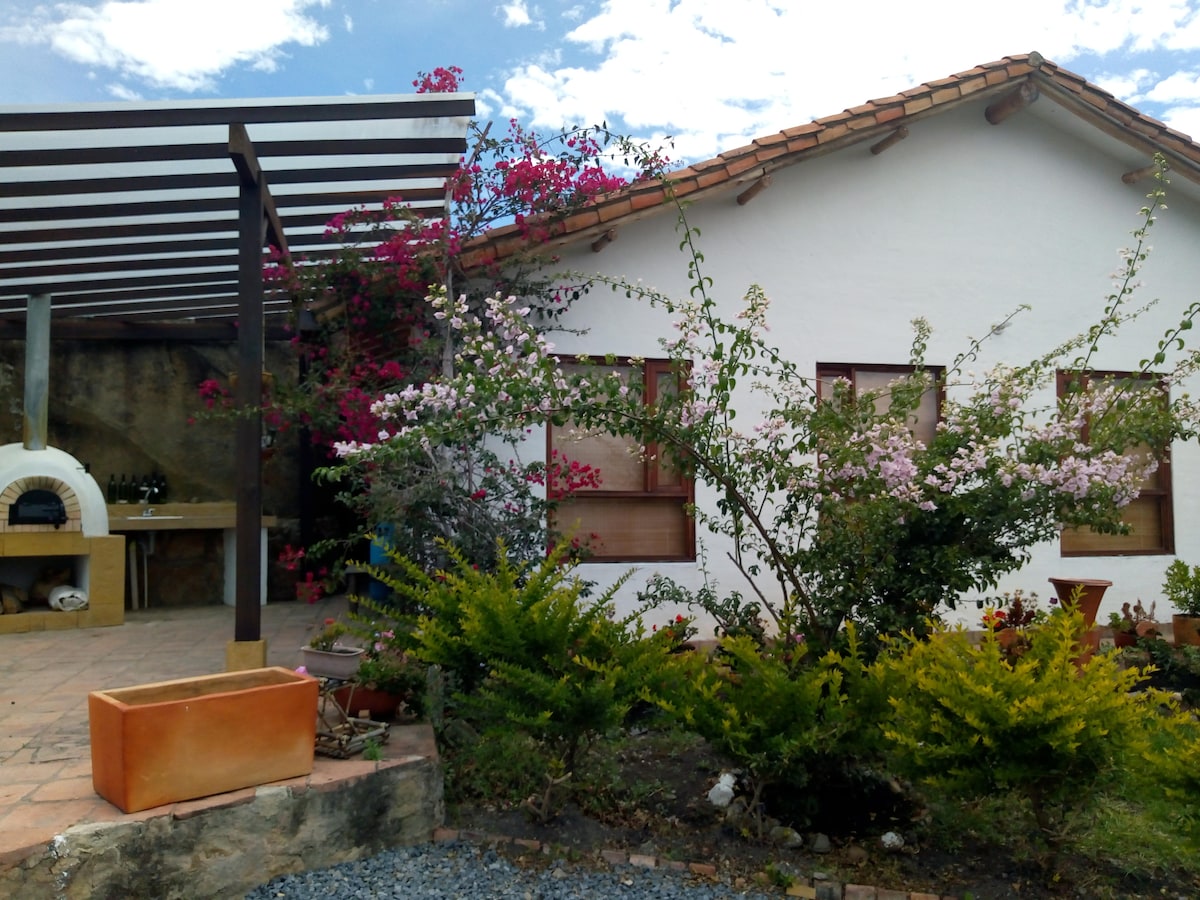 A charming exterior view of the house is framed by colorful flowering plants, enhancing the natural setting. A shaded patio with a wood-fired oven is visible, alongside an orange planter. The sunlight illuminates the white walls and a clear blue sky can be seen overhead.