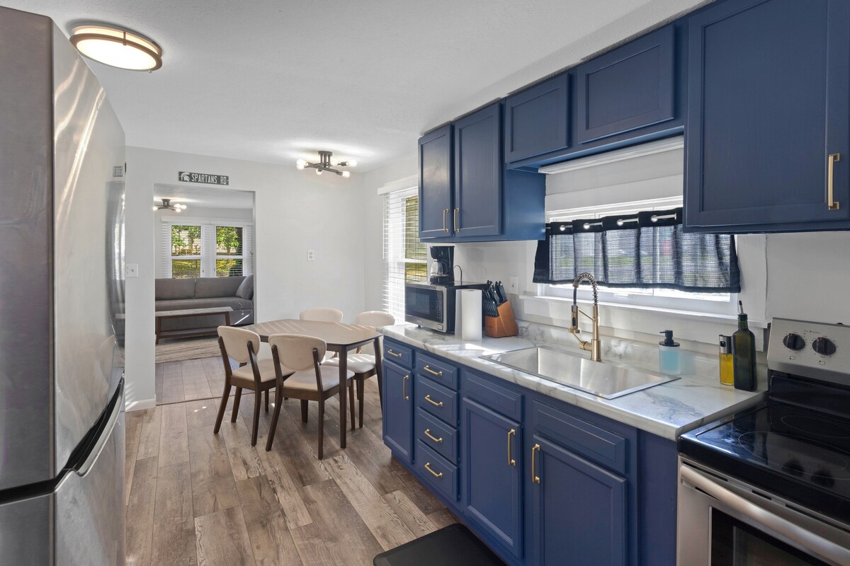 The kitchen features navy blue cabinetry and stainless steel appliances. A spacious countertop provides ample prep space, while a dining area with a round table and four chairs is located adjacent. Natural light filters through the window, illuminating the modern design.