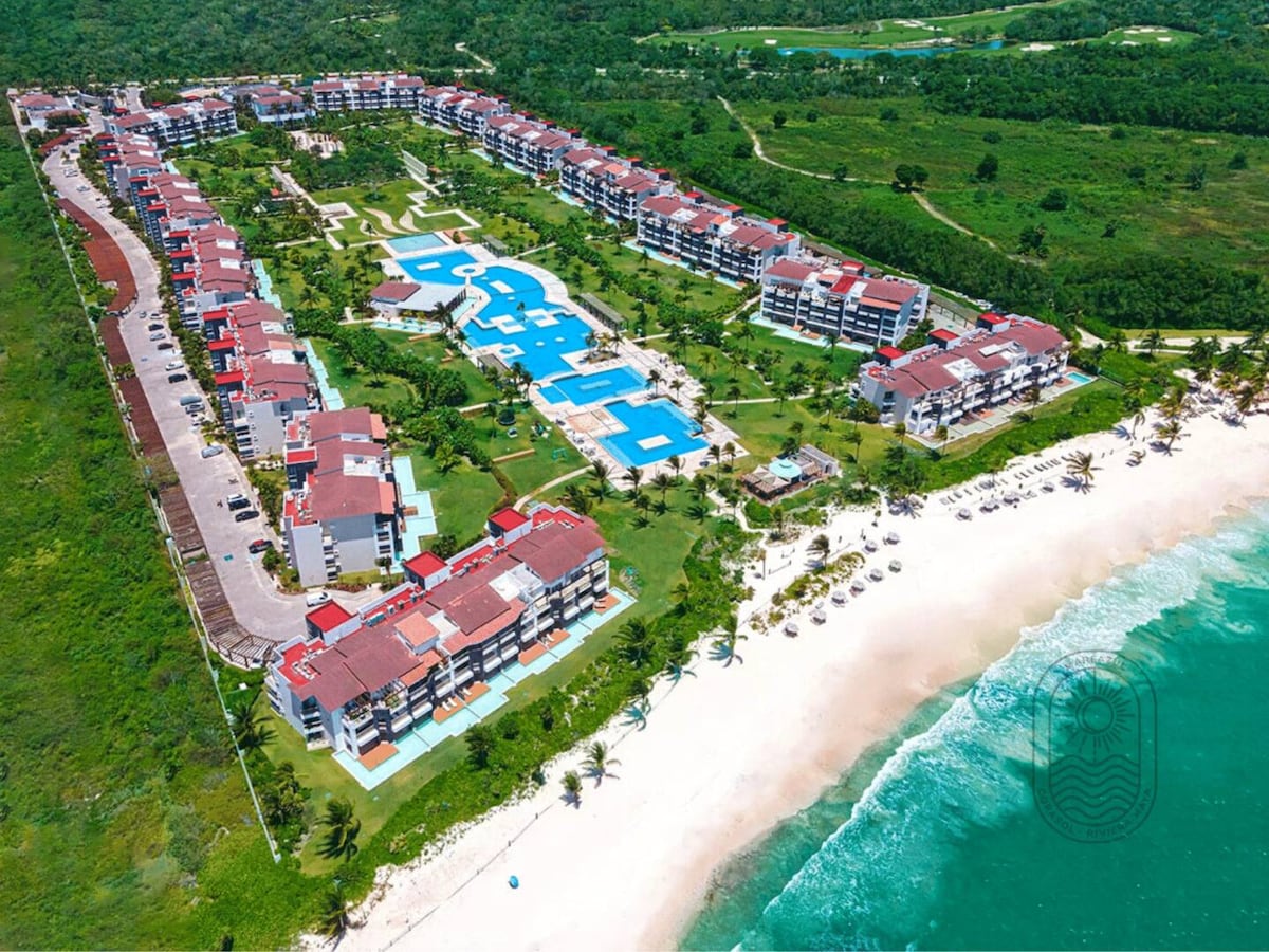 An aerial view captures the expansive Mareazul complex, displaying multiple buildings surrounded by lush greenery, turquoise pools, and a private beach. The layout reveals pathways lined with palm trees, leading to sandy shores and lounge areas by the water's edge.