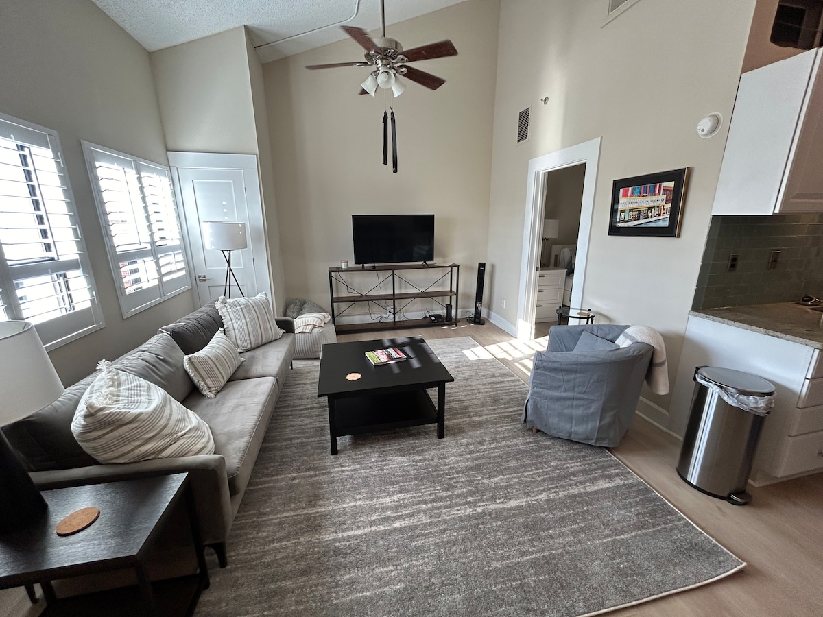 A spacious living area is showcased with a comfortable sofa and lounge chair, arranged around a dark coffee table. A flat-screen TV is positioned on a contemporary media console. Natural light filters through the large windows with plantation shutters, illuminating the neutral decor and plush area rug.
