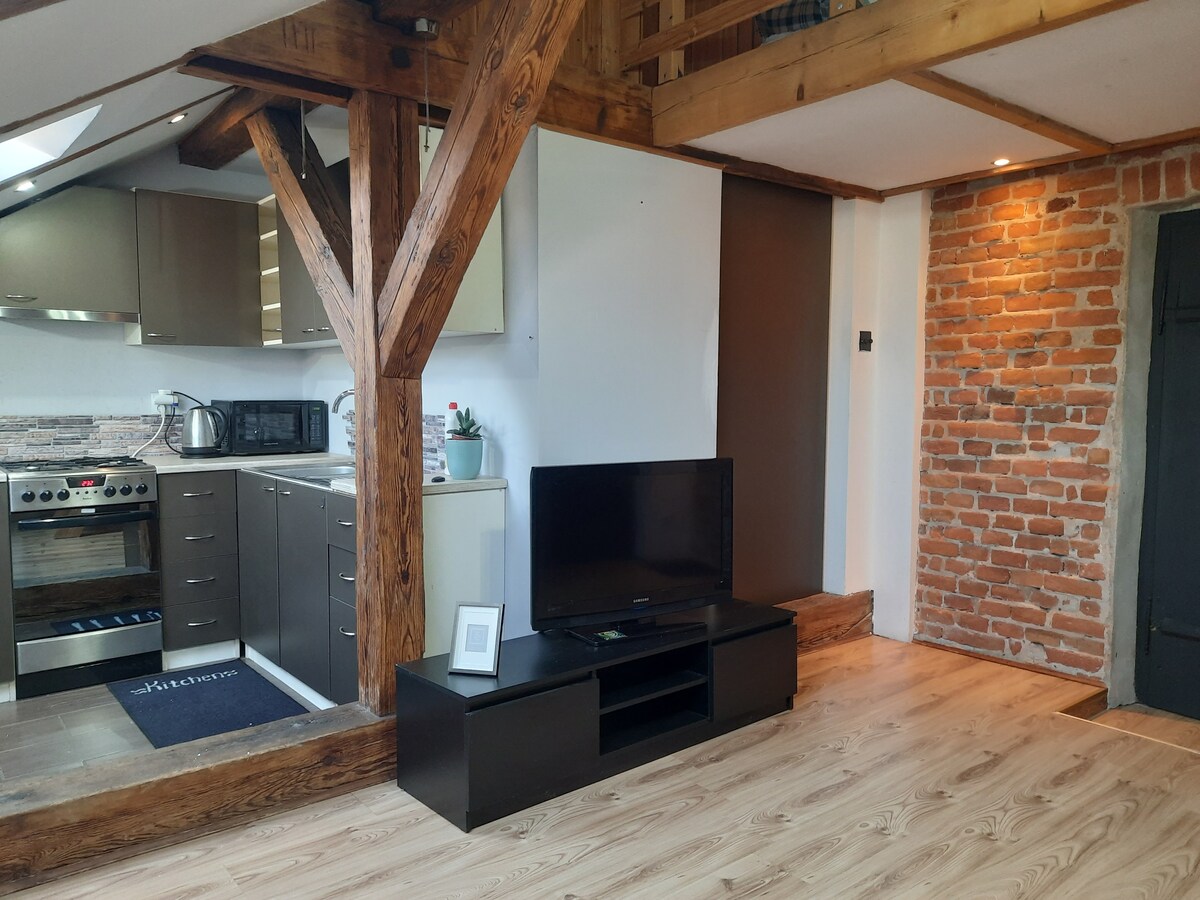 A cozy space with exposed wooden beams and a rustic brick wall is visible. A modern kitchen with stainless steel appliances, including a stove and microwave, is featured. A television is positioned on a low black media unit, creating a blend of contemporary and traditional elements.