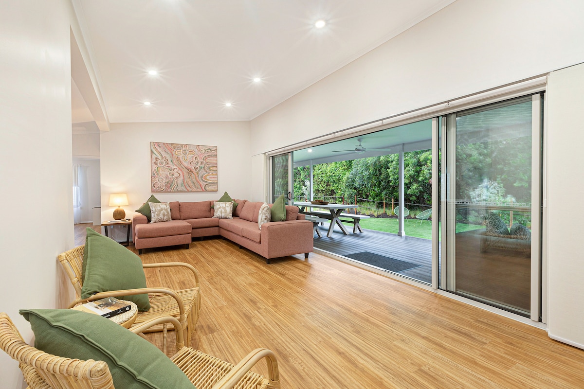A spacious living area features a light-coloured floor and a comfortable pink sectional sofa with green decorative cushions. Large sliding glass doors open to a deck area, allowing for natural light and a view of the lush greenery outside. Two wicker chairs are positioned nearby.