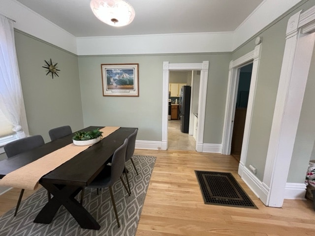 A dining area features a dark wood table surrounded by chairs, complemented by a neutral-colored rug. A decorative plant rests in the center of the table. Soft, natural light filters through sheer curtains, illuminating the light green walls and contrasting with the white trim.
