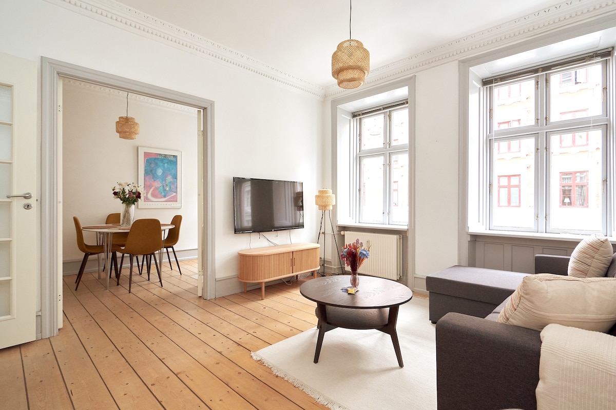 An open living area features a comfortable gray sofa, round coffee table, and a large television mounted on the wall. Natural light streams through multiple windows, illuminating the space. A dining area with modern chairs and a vase of flowers is visible in the background.