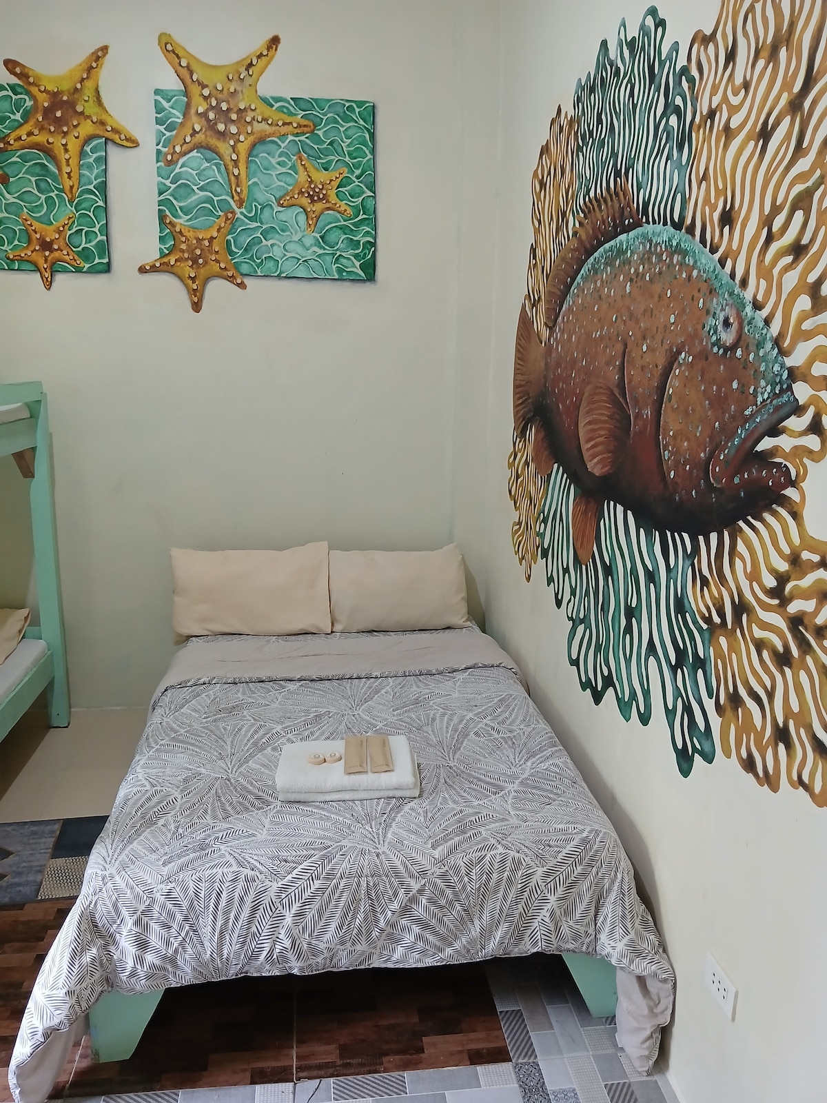 A colorful mural depicting a fish surrounded by coral covers one wall. A well-made bed is adorned with a neutral-toned blanket and two cream-colored pillows. Towels are neatly arranged on the bed, while decorative starfish artwork adds a playful touch to the room.