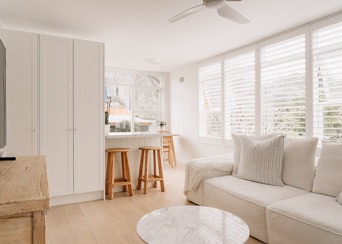 A bright living area is highlighted by white walls and natural light filtering through plantation shutters. A cozy sectional sofa sits beside a round marble coffee table, while two wooden stools accompany a small dining table. Light oak flooring enhances the inviting ambiance.