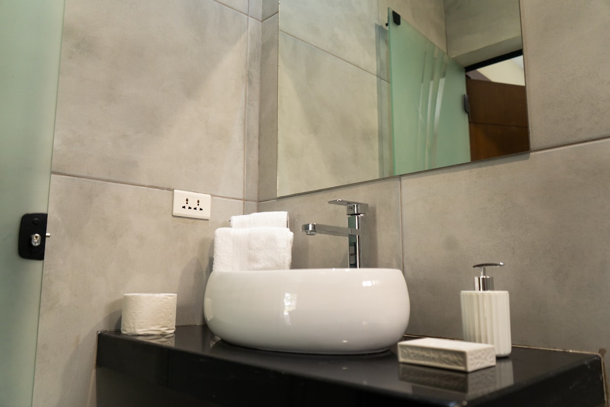 A modern bathroom features a sleek sink with a smooth circular basin. Fresh white towels are neatly arranged beside it. A minimalist mirror is mounted on the wall above, reflecting the neutral tones of the surrounding tile and countertop.