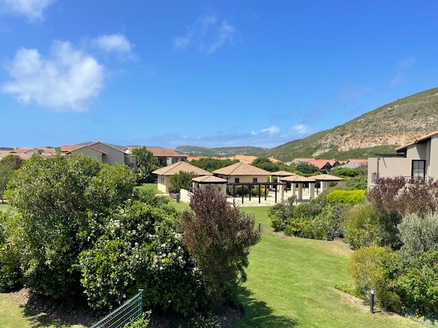 Lush greenery surrounds a well-maintained complex featuring modern architecture and a variety of outdoor spaces. The landscape is interspersed with trees and shrubs, with a clear blue sky above and gentle hills in the background.