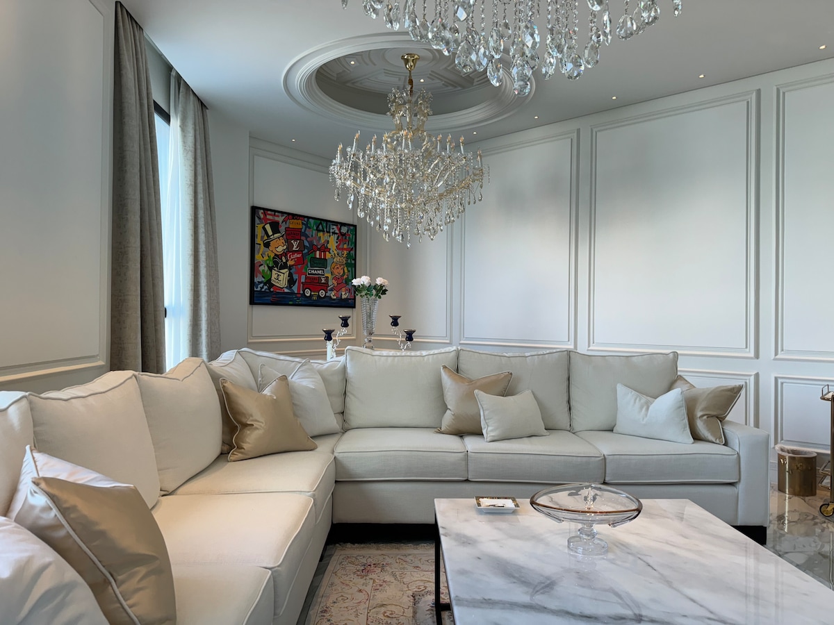A spacious living area features a large white sectional sofa adorned with decorative cushions. A modern chandelier hangs from the ceiling, illuminating the room. A marble coffee table sits centrally, complemented by an intricate area rug and artistic wall decor.