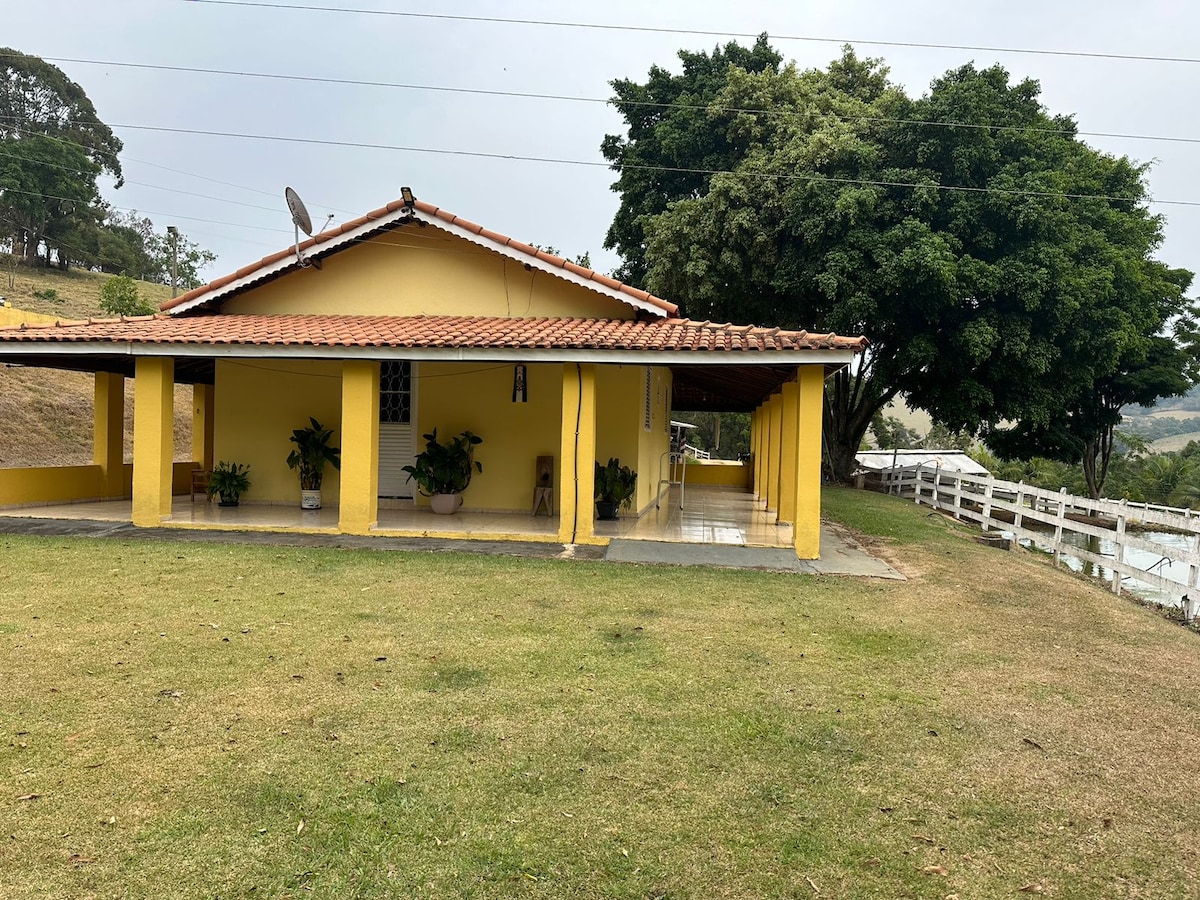 A yellow single-story house is seen from the side, featuring a covered porch supported by wooden posts. Potted plants adorn the porch area, and a vast lawn extends towards a tree line in the background. A white fence runs parallel to the property.