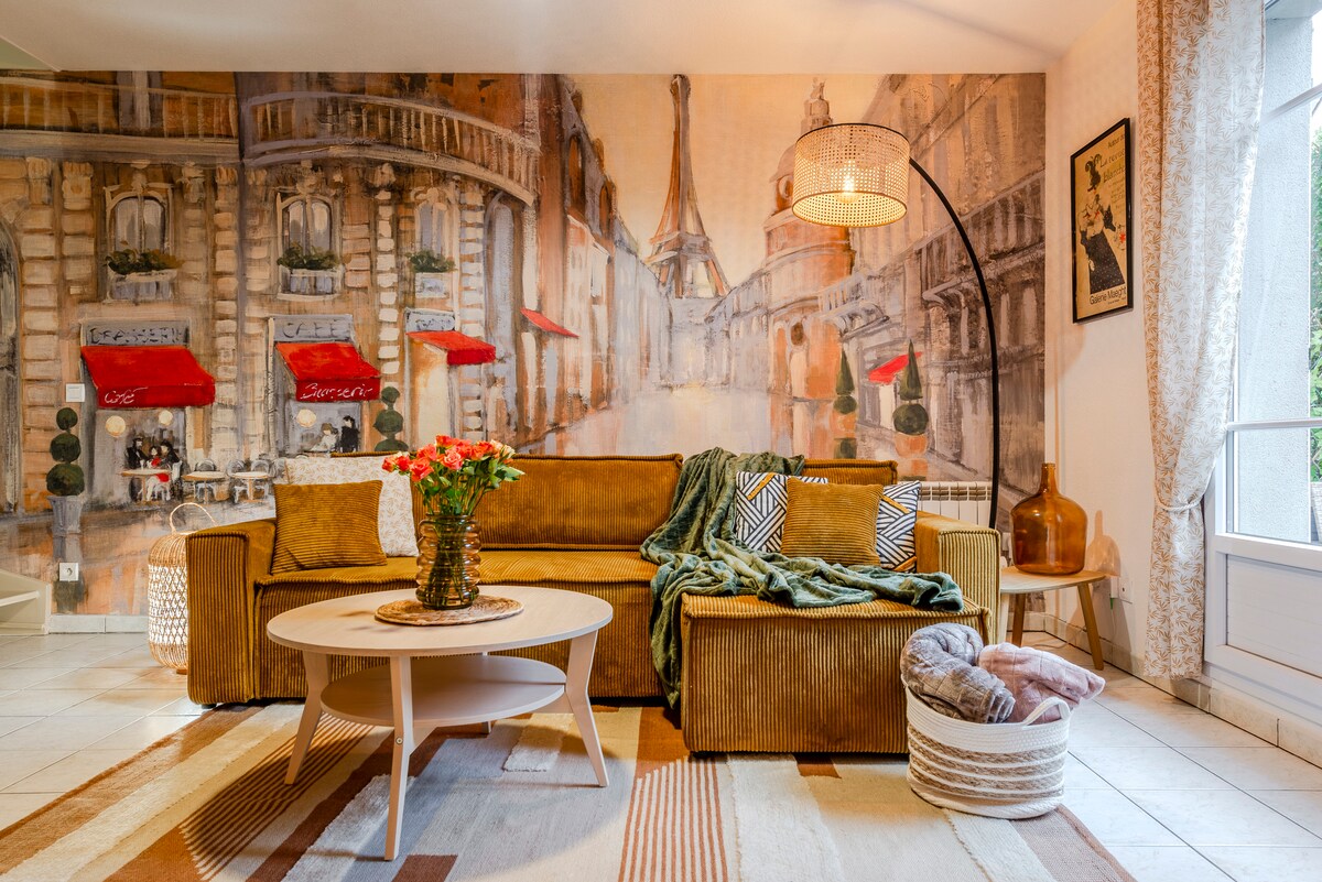 A living room features a large, comfortable sofa adorned with patterned cushions and a textured throw. A round coffee table centers the space, complemented by an artistic wall mural depicting a Parisian street scene. Natural light fills the room through large windows.