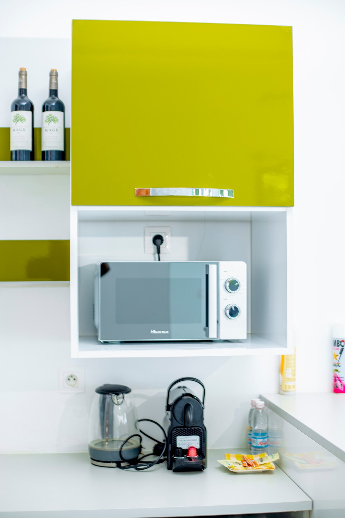 A modern kitchen area is showcased with a vibrant green cabinet overhead. A microwave is positioned below, accompanied by a kettle and a coffee maker. Bottles of wine are displayed on the shelf above, adding a touch of elegance to the space.