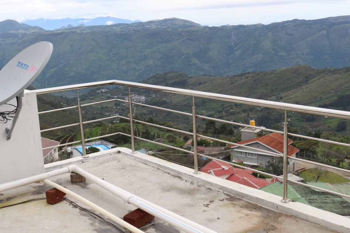 A balcony offers a panoramic view of the surrounding mountains and valleys. The railing is made of stainless steel, contributing to a modern aesthetic. The concrete floor is visible, along with plumbing elements in the foreground, enhancing the outdoor space's utility.