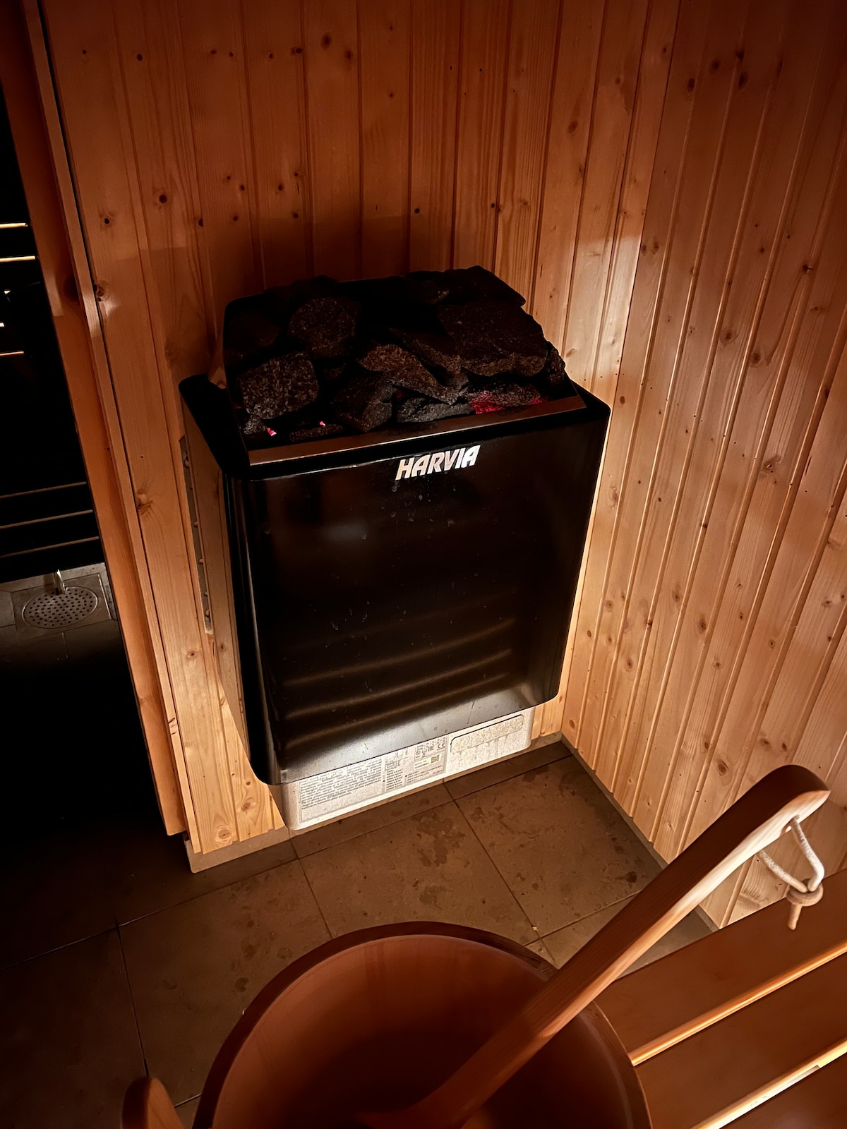 A modern sauna heater is mounted on a wooden wall, featuring a black exterior and a top filled with stones. Dark wooden paneling surrounds the heater, creating a warm and serene atmosphere. A wooden bucket and ladle are visible on the tiled floor.