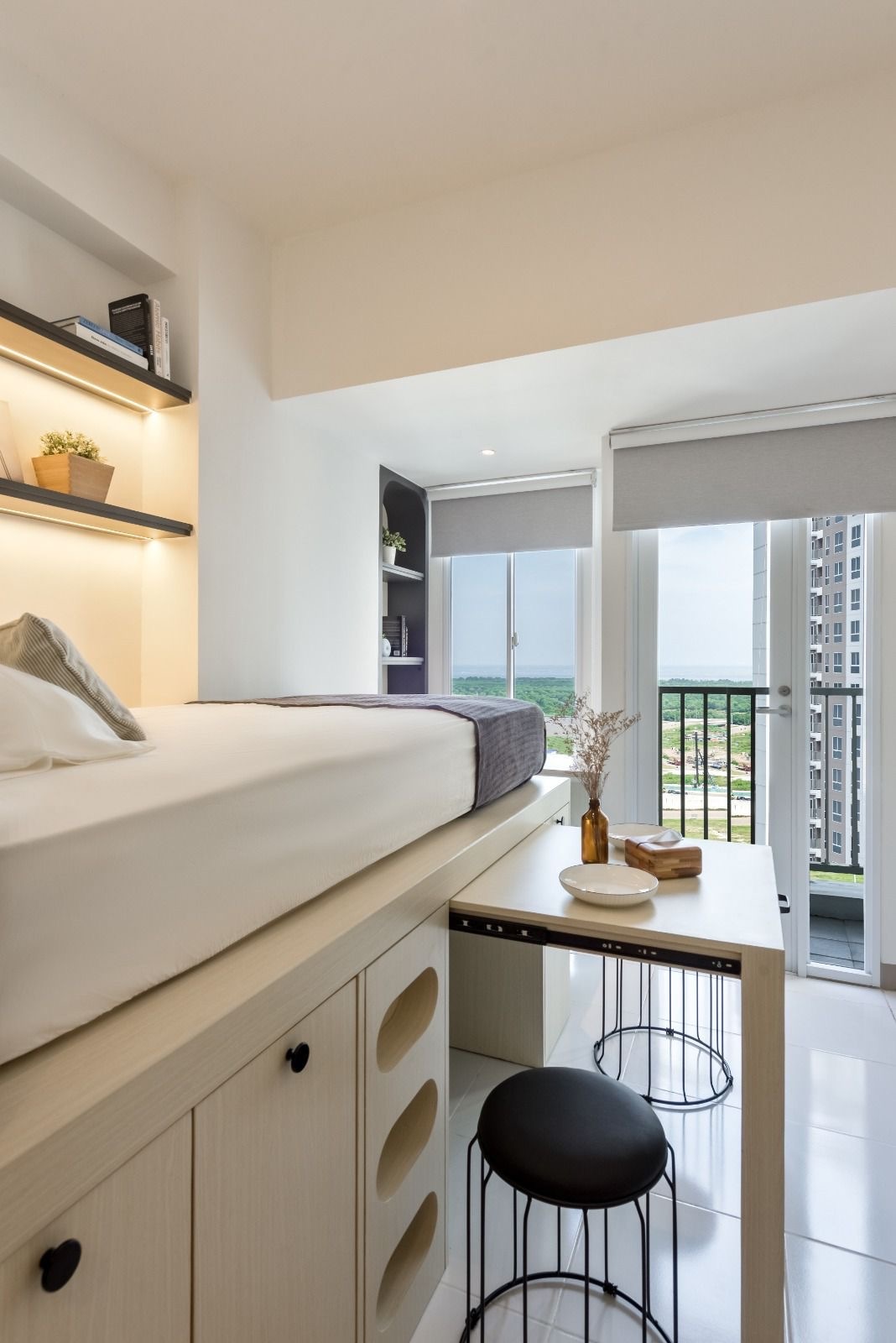A modern studio interior is displayed, featuring a spacious elevated bed with soft linens. A small desk is positioned nearby, complemented by a black stool. Natural light fills the area through large windows with roller shades, providing scenic views of the outdoors.