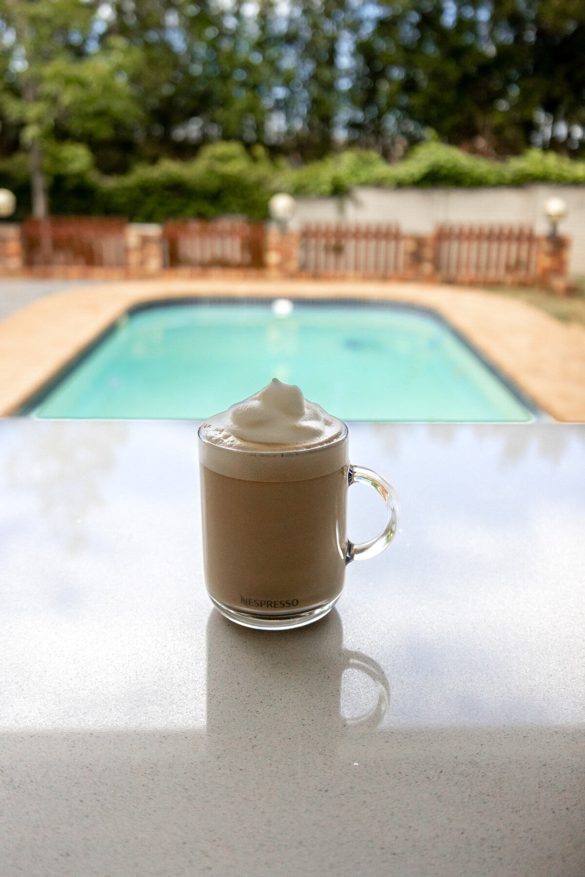 A glass of coffee topped with whipped cream is placed on a smooth countertop. In the background, a clear blue pool is visible, surrounded by a patio area with wooden fencing and landscaped greenery.