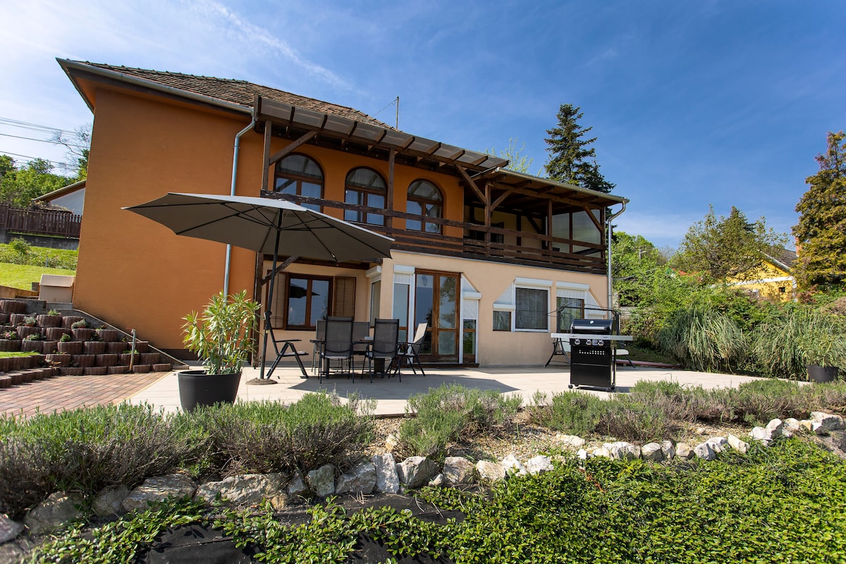 A spacious outdoor terrace is shown, featuring a dining set with dark chairs under a large umbrella. The house, with its warm-colored exterior, is complemented by a well-kept garden and a barbecue grill. Lush greenery surrounds the area, enhancing the natural setting.