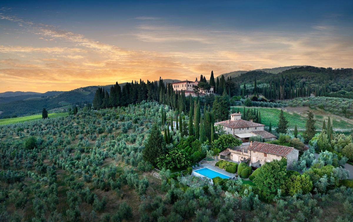 A panoramic view of a villa surrounded by lush greenery and rolling hills. The property features a private pool, and mature trees and shrubs enhance the scenic landscape, providing a serene retreat. The sky displays soft, warm hues as the sun begins to set.