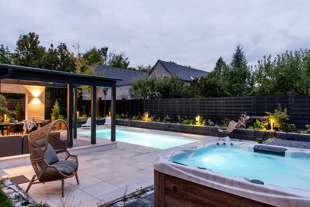 An outdoor space features a well-lit swimming pool alongside a jacuzzi. Modern seating arrangements are provided under a pergola, surrounded by greenery and privacy fencing. Soft ambient lights enhance the tranquil atmosphere, creating an inviting area for relaxation.