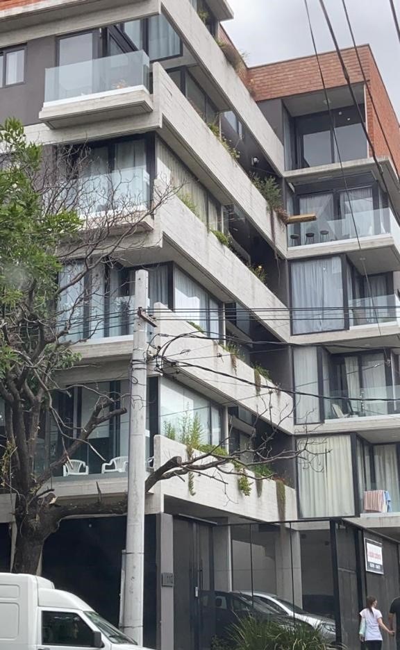 A modern multi-story building is depicted, featuring balconies with glass railings. Natural greenery is integrated throughout the structure, enhancing its aesthetic appeal. Large windows provide ample natural light, creating a bright and airy atmosphere. The facade combines concrete and glass elements.