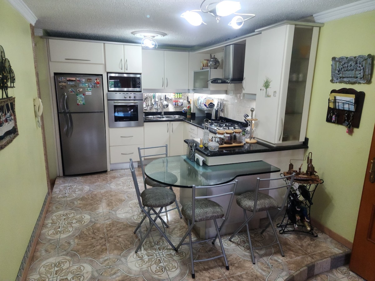 A modern kitchen is presented, featuring sleek cabinetry and stainless-steel appliances. A round dining table with four chairs is situated in the center. Bright overhead lighting illuminates the space, while decorative tile flooring adds texture and visual interest.
