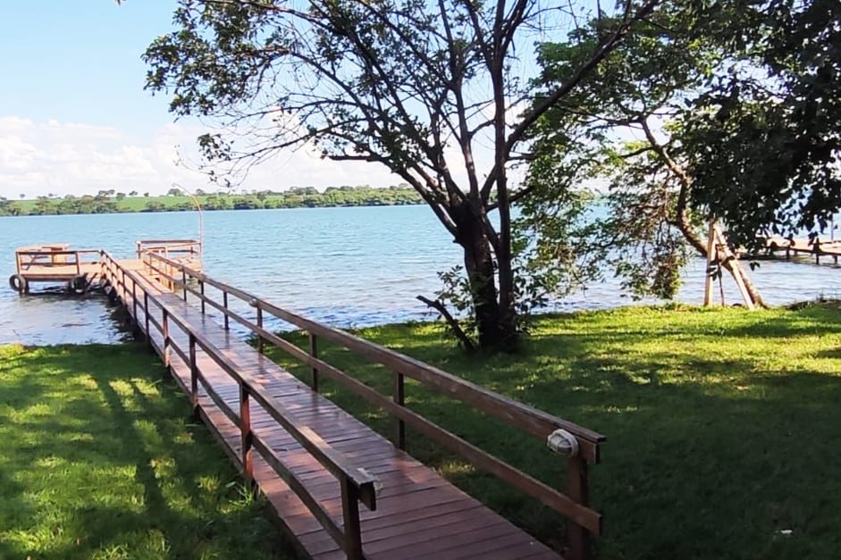 A wooden walkway leads towards the water, framed by lush green grass and trees. The tranquil lake is visible in the background, reflecting a clear blue sky with scattered clouds.