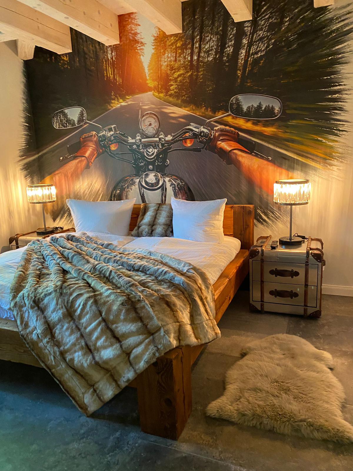 A spacious bedroom is depicted with a wooden bed adorned with white pillows and a plush, neutral blanket. On either side of the bed, stylish lamps provide soft lighting. A striking mural of a motorcycle on a winding road adds a dynamic backdrop to the serene space.