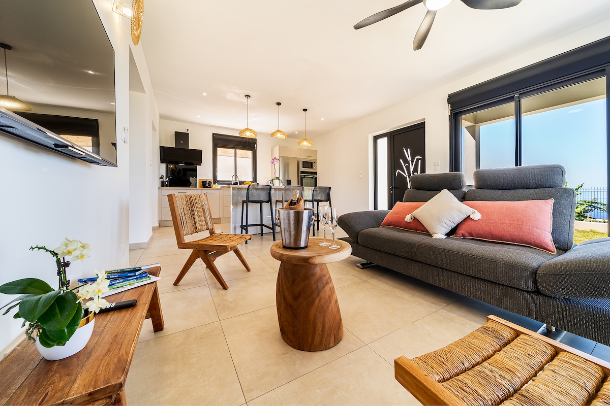 An open-plan living area is highlighted, featuring a comfortable sectional sofa with accent pillows. A round wooden table is positioned in the center, surrounded by contemporary chairs. A flat-screen television is mounted on a wall, while large windows bring in natural light, revealing outdoor views.