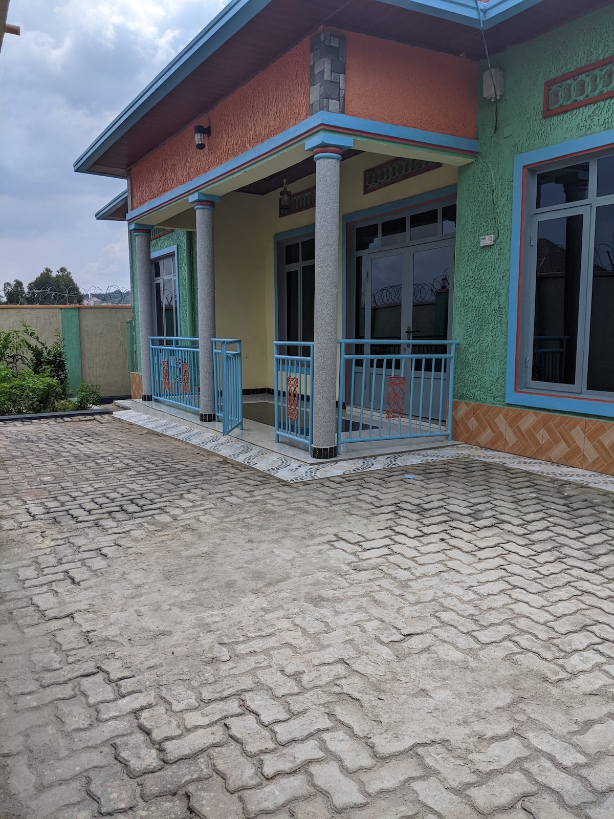 A front porch is seen with a colorful exterior, showcasing blue railings and large windows. Light-colored paving stones form a pathway leading to the entrance, bordered by green grass and shrubs. The sky is partly cloudy, adding a serene backdrop.