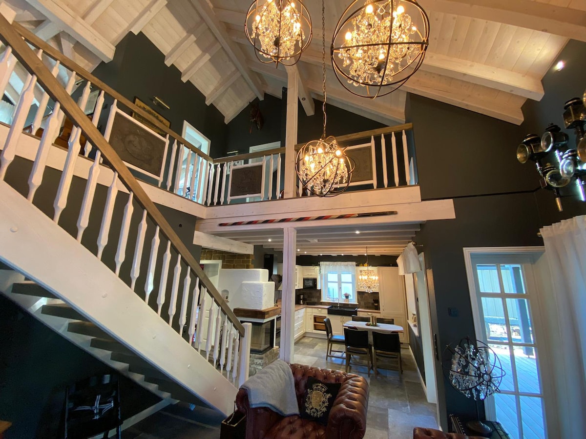 A spacious living area showcases high ceilings with wooden beams and two decorative chandeliers. An open staircase leads to a loft area, while a leather sofa is positioned near a dining table visible in the background. Large windows allow natural light to illuminate the space.