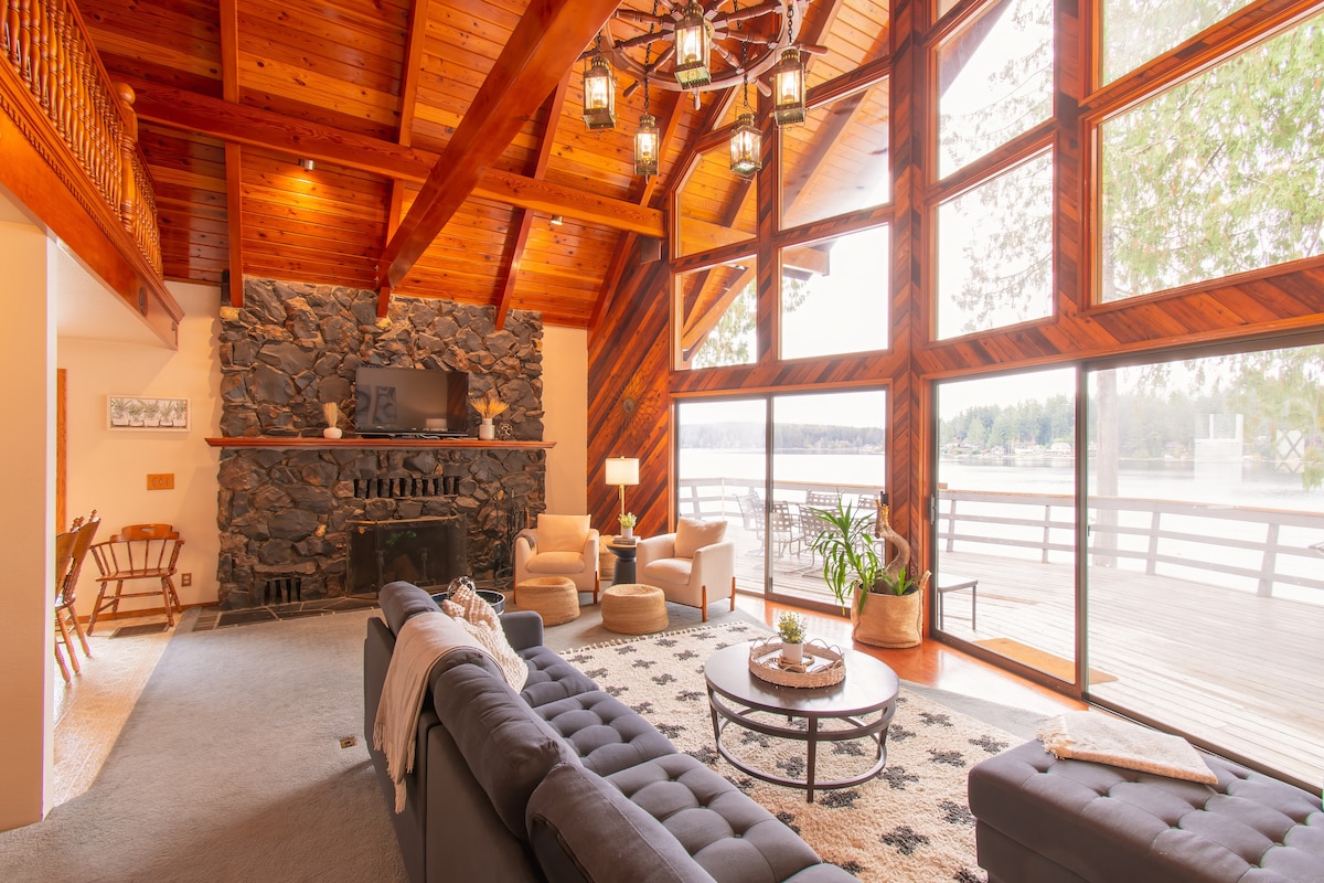 A spacious great room is showcased with vaulted wooden ceilings and large windows offering views of the lake. A stone fireplace serves as a focal point, while a comfortable seating area includes a dark couch and side chairs. Natural light floods the space, enhancing its inviting ambiance.