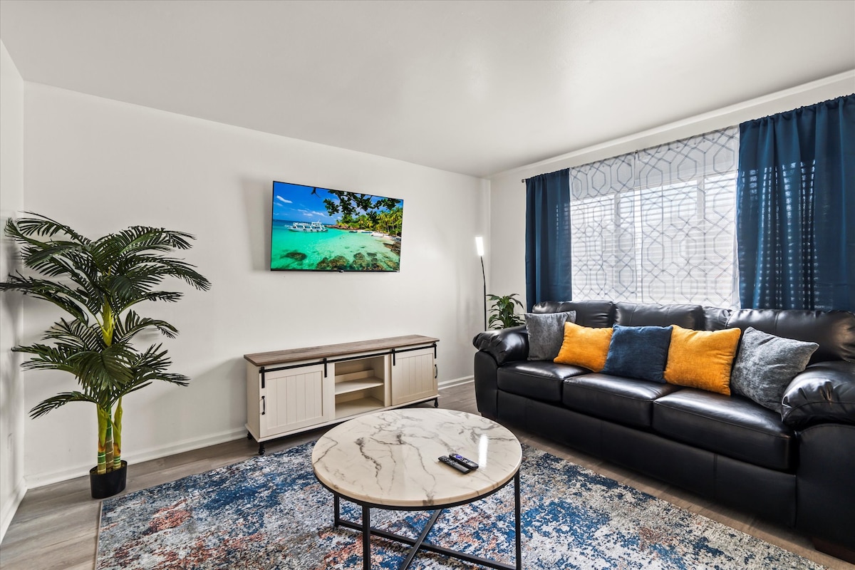 A comfortable living area features a black sofa adorned with orange pillows. A coffee table with a marble top sits in the center. A television mounted on the wall displays a scenic beach view, while large windows dressed with dark curtains allow natural light to enter. A decorative plant adds a touch of greenery.