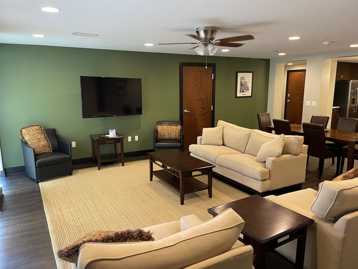 A spacious living area features two beige sofas and two black armchairs arranged around a dark wooden coffee table. A flat-screen television is mounted on a green accent wall, while a dining area with a large table is visible in the background, enhancing the comfort of the space.