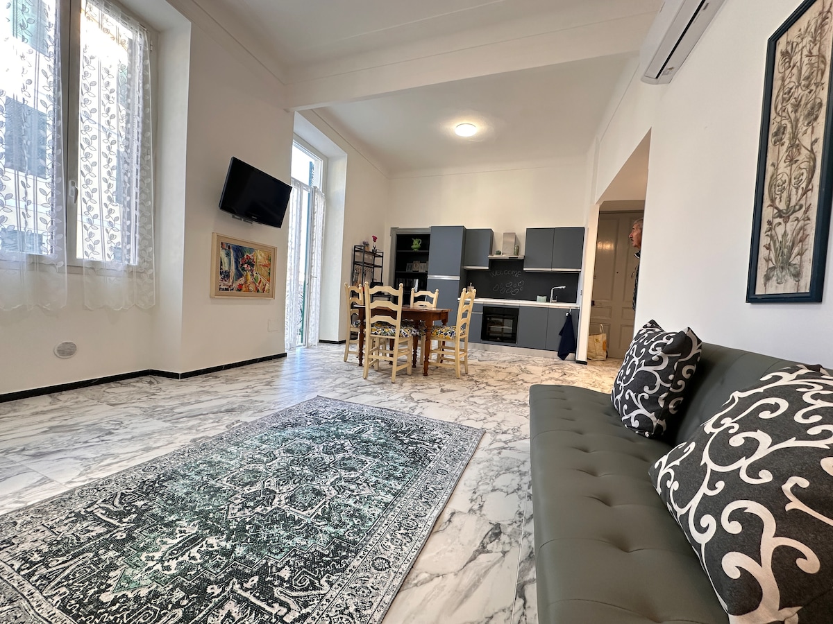 A spacious living area is visible, featuring a sofa adorned with decorative cushions, a dining table for five, and a well-equipped kitchen. Natural light enters through large windows, highlighting the elegant marble flooring and tasteful artwork on the walls.