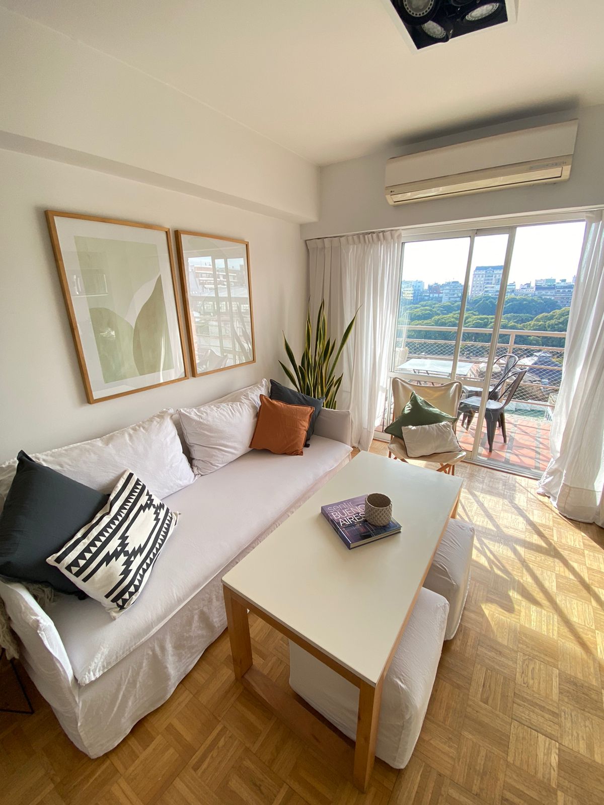 A bright living area features a light-colored sofa adorned with coordinating cushions. A small coffee table sits nearby, holding a book. Natural light fills the space through large glass doors that lead to a balcony with city views, enhancing the open atmosphere.