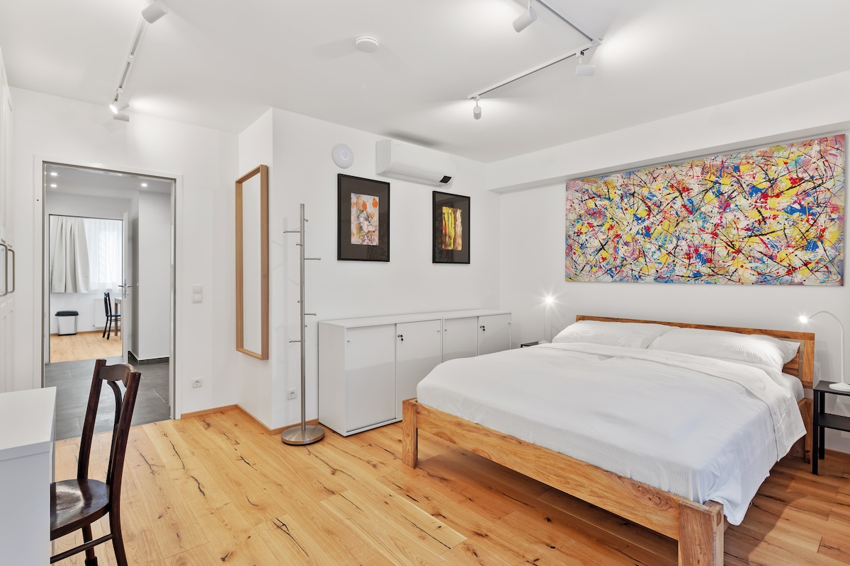 A spacious bedroom features a warm wooden floor and a large bed with crisp white linens. Colorful abstract art is displayed above the bed, adding a vibrant touch to the minimalist decor. A white sideboard provides additional storage, and natural light enters through the door leading to another room.